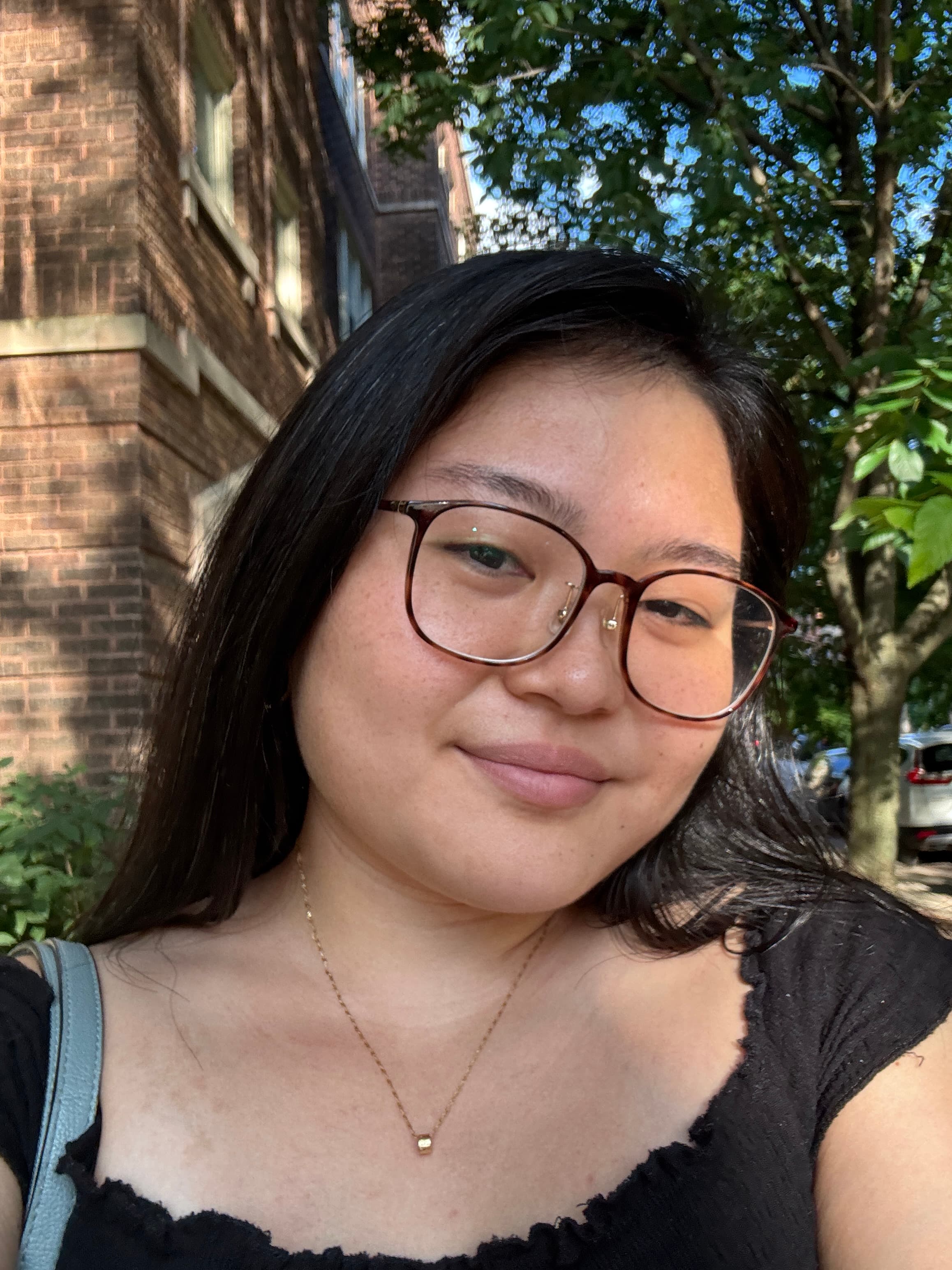 Selfie of Reina smiling. They are wearing brown square-ish glasses and long black hair.